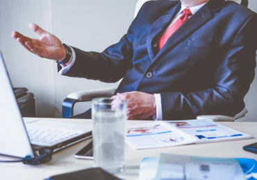 Man sitting on a chair talking with hand outstretched, over a table with a glass of water, note book and papers ( Breach Of Contract_Asia_Law_Network)