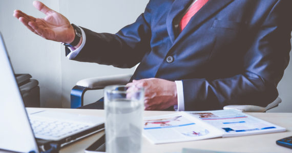 Man sitting on a chair talking with hand outstretched, over a table with a glass of water, note book and papers ( Breach Of Contract_Asia_Law_Network)