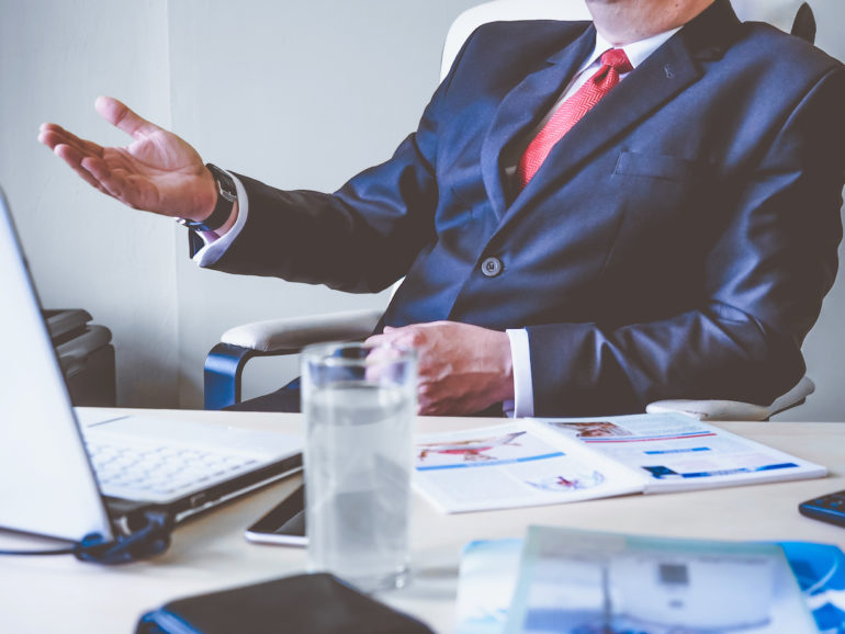 Man sitting on a chair talking with hand outstretched, over a table with a glass of water, note book and papers ( Breach Of Contract_Asia_Law_Network)